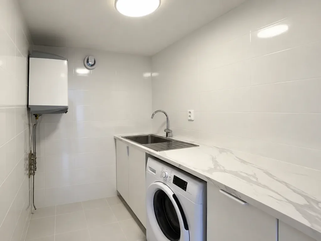 Nyrenoverad tvättstuga i Karlstad med vit inredning, tvättmaskin, bänkskiva med diskho och ljusa kaklade väggar.